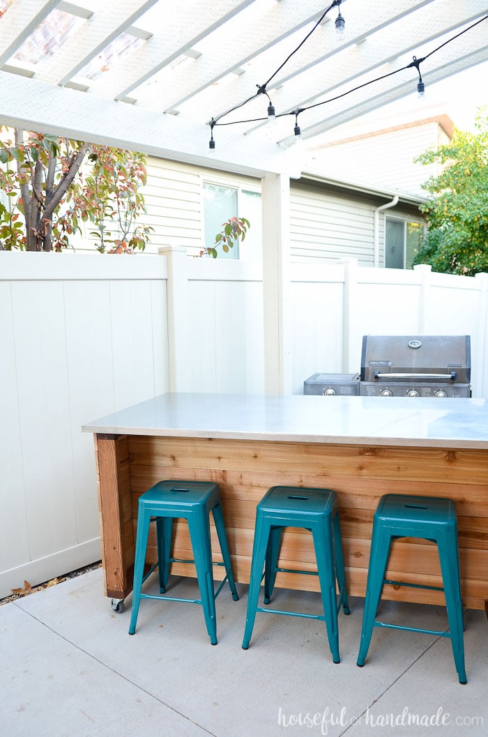 DIY outdoor bar cart with a large countertop, wood plank base, locking casters, and teal bar stools under a covered backyard patio with string lights.