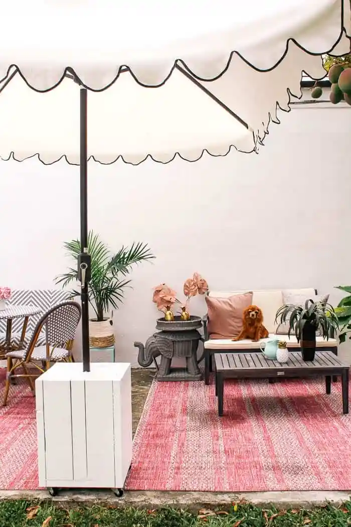 White rectangular outdoor umbrella stand with a patio umbrella mounted through the center, positioned beside a seating area with a sofa, coffee table, and pink outdoor rug.