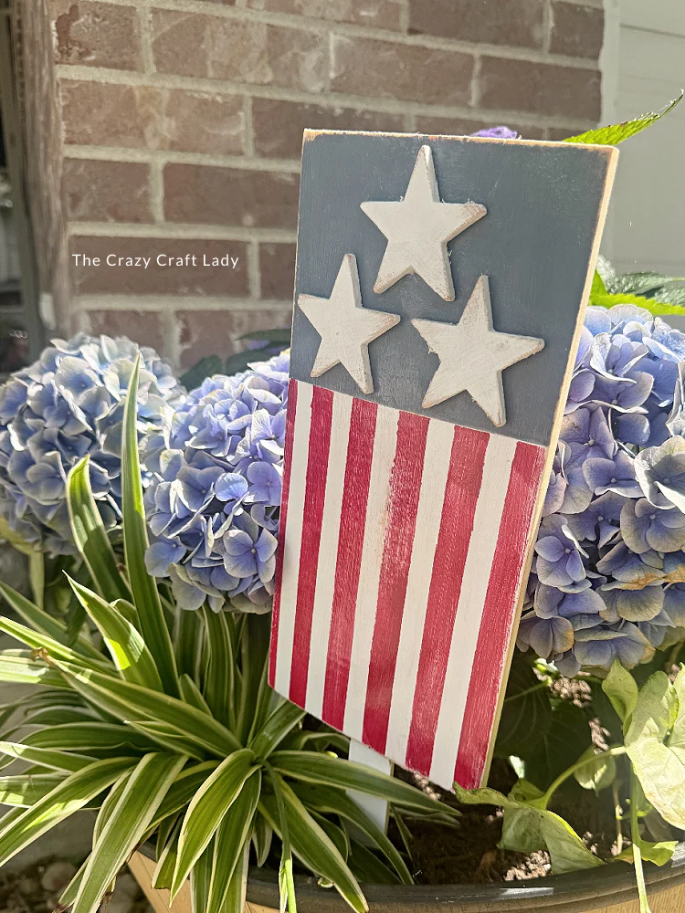 Patriotic garden stake featuring a hand-painted wooden flag design with red-and-white stripes and three white stars on a blue background