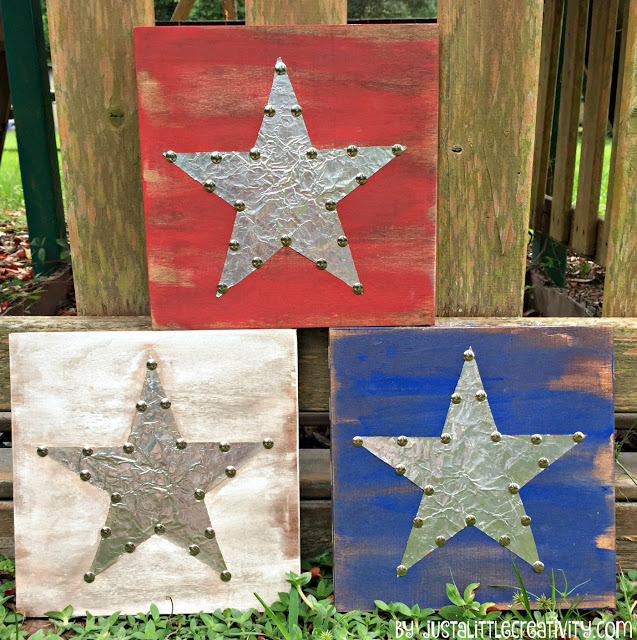 Three square wooden plaques leaning against a weathered wooden railing outdoors. Each plaque features a large metallic silver star in the center, outlined with decorative metal tacks.