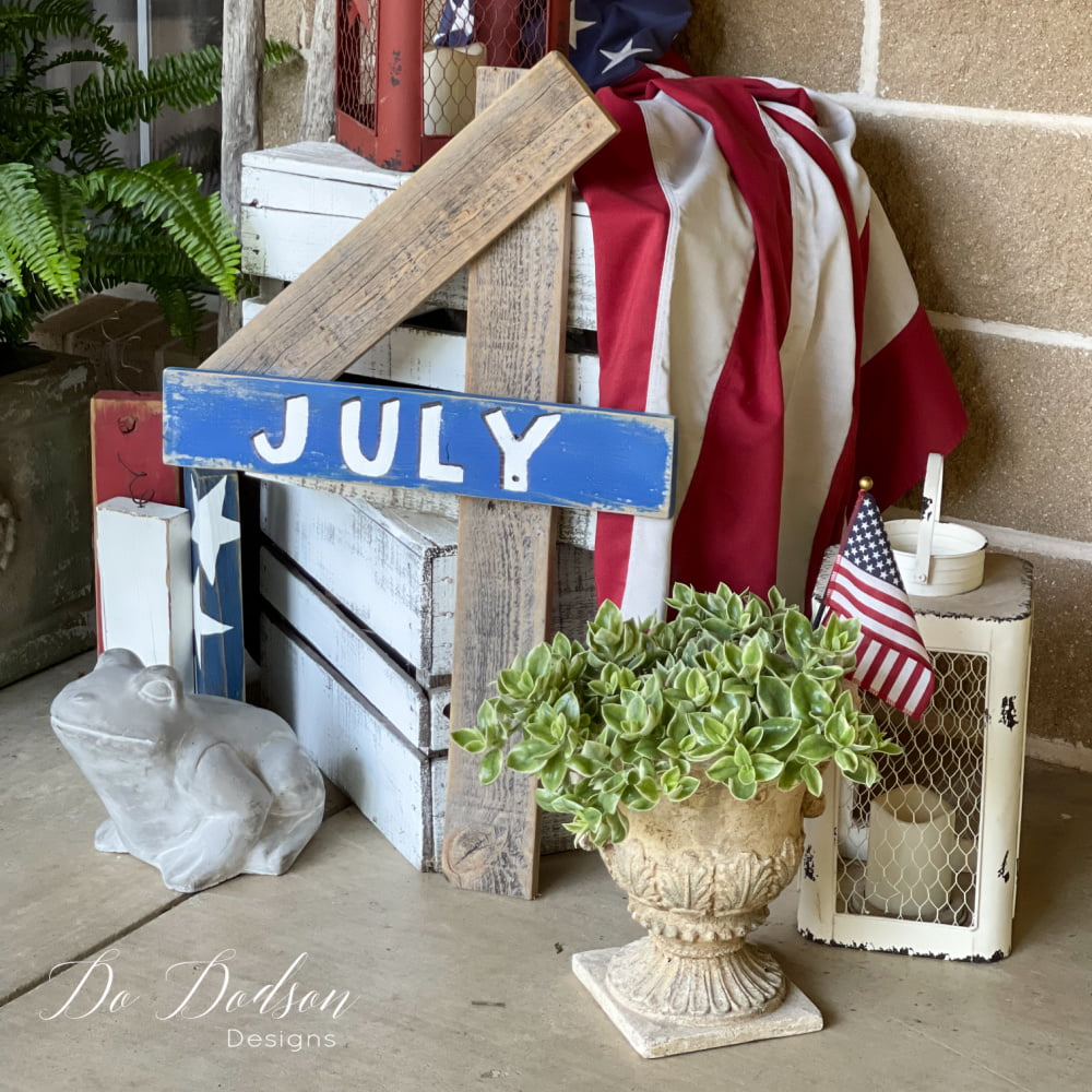 Rustic patriotic porch display featuring a weathered wooden ‘July’ sign with red, white, and blue accents.
