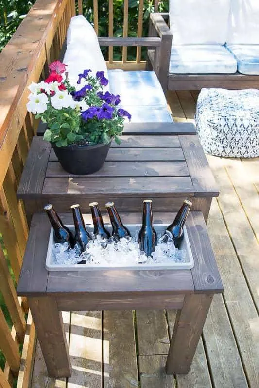 DIY outdoor side table with a built-in cooler shelf holds bottles chilled on ice, while the upper tabletop displays a colorful potted flower arrangement.