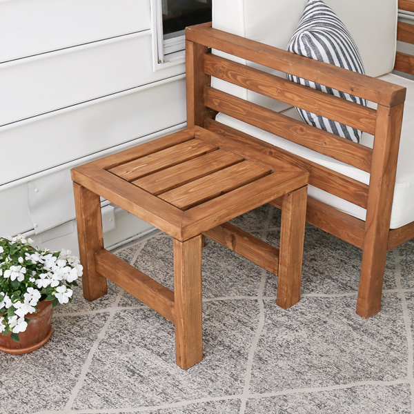 Wooden outdoor side table with a slatted top and square legs positioned beside a patio chair on a porch.