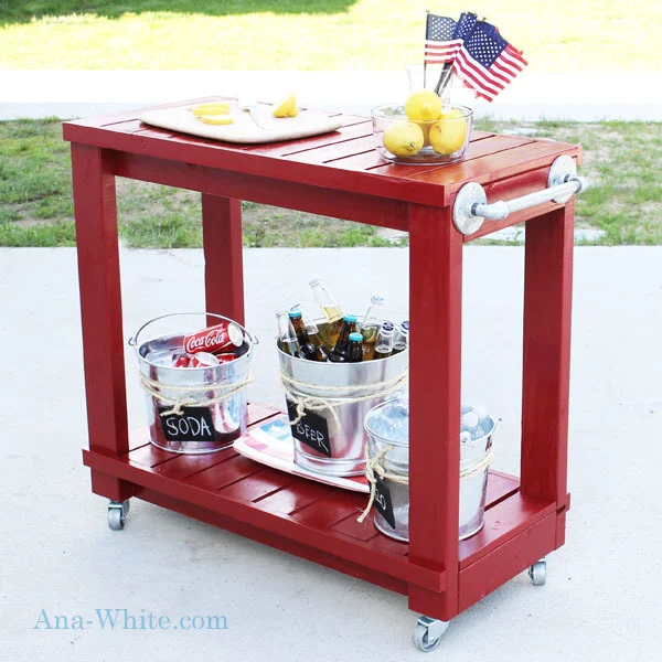 Red rolling outdoor cart with pipe-style towel handles, lower storage shelf, metal drink buckets, and small American flags on a patio.