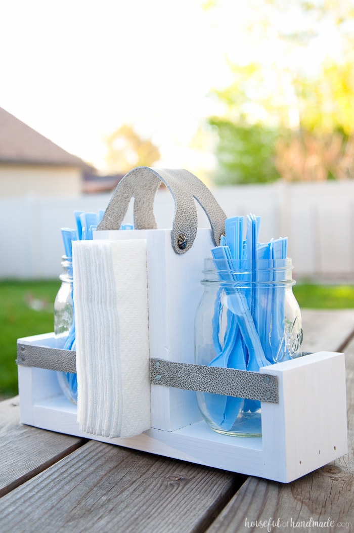 White wooden utensil caddy with straps holding mason jars filled with blue plastic cutlery and napkins on an outdoor table.