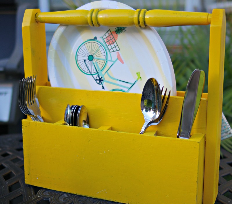 Yellow wooden utensil caddy holding paper plates, forks, spoons, and knives on an outdoor table.