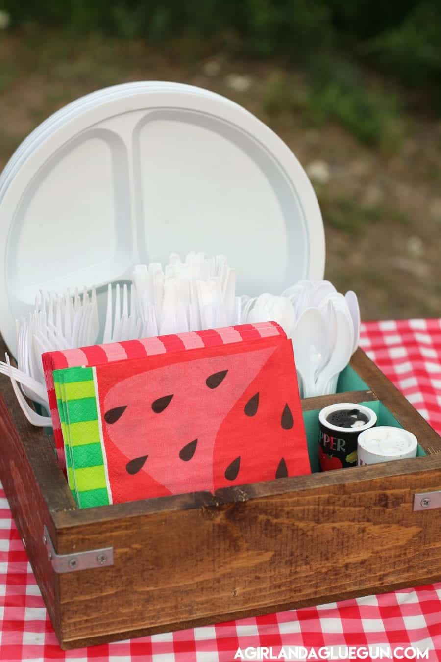 Wooden picnic caddy holding paper plates, plastic utensils, watermelon napkins, and condiments on a red gingham tablecloth.