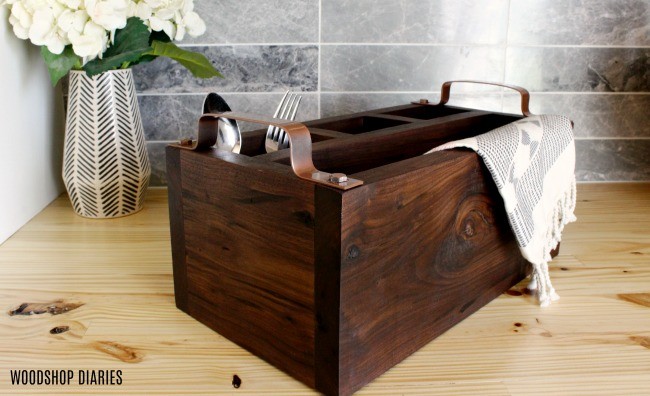 Dark stained wooden utensil caddy with metal handles holding utensils and a towel on a wood countertop.